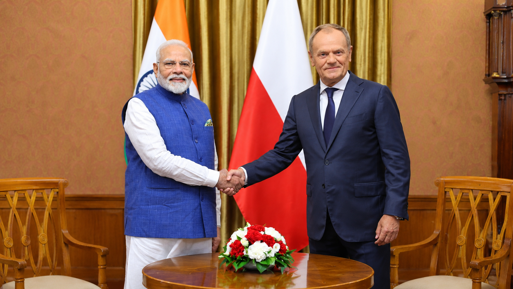Prime Minister Narendra Modi and Polish Prime Minister Donald Tusk shaking hands during a diplomatic meeting, with Indian and Polish national flags in the background and a flower arrangement on a table.